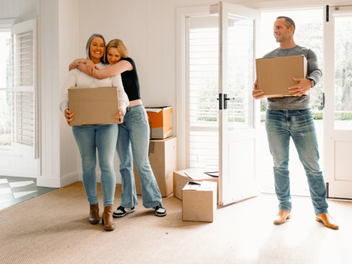 Happy couple carrying cardboard boxes while teenage girl hugs mum from behind inside home - Australian Stock Image