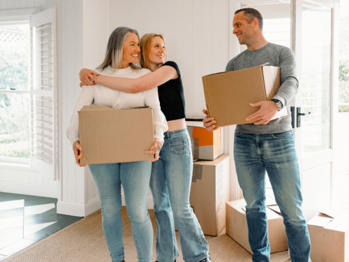 Happy couple carrying cardboard boxes while teenage girl hugs mum from behind inside home - Australian Stock Image