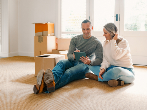 Happy couple seated on carpeted floor of their new home holding a tablet device - Australian Stock Image