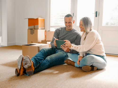 Happy couple seated on carpeted floor of their new home holding a tablet device - Australian Stock Image