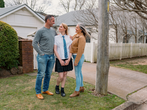 Happy couple stands closely with teenage daughter in school uniform outside of their new home - Australian Stock Image