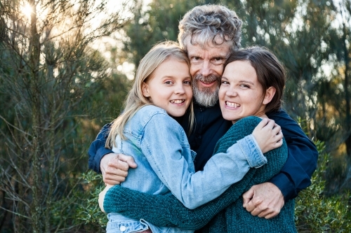 Happy dad with his two teenage daughters hug together outside - Australian Stock Image