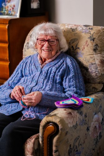 happy elderly woman sitting on armchair crocheting blanket square for great grandchild - Australian Stock Image