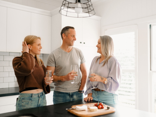 Happy family gathered around the kitchen countertop sharing food and adults drinking wine - Australian Stock Image