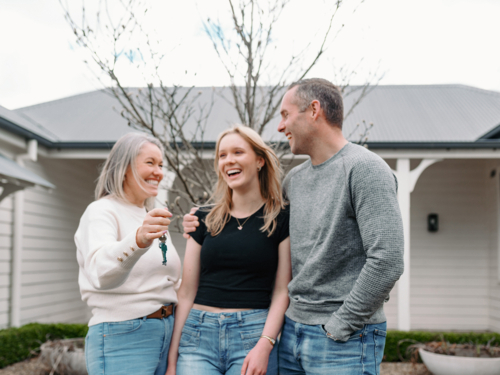 Happy family standing in front yard with mum holding out the key of their new home - Australian Stock Image