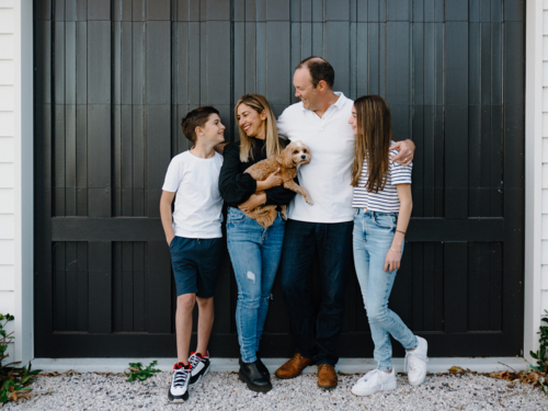 Happy family standing outside in front of black garage door with pet dog - Australian Stock Image