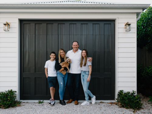 Happy family standing outside in front of black garage door with pet dog - Australian Stock Image