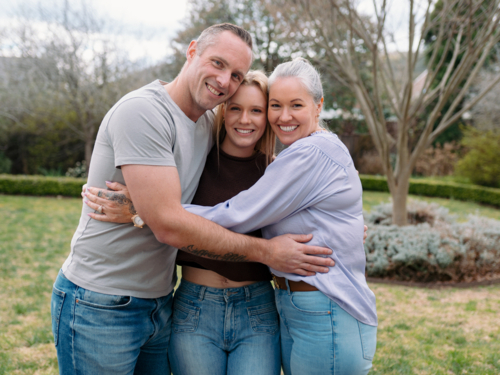 Happy family stands closely hugging each other in the garden of their new home - Australian Stock Image