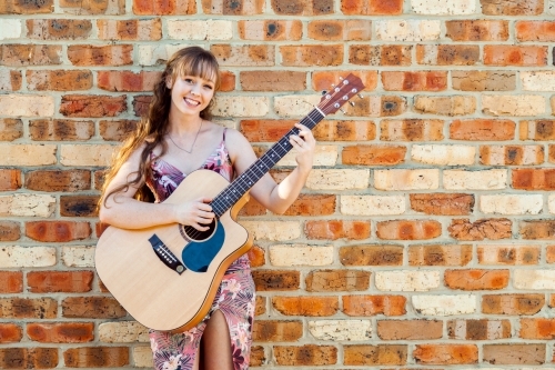 Happy female musician with guitar against brick wall - Australian Stock Image