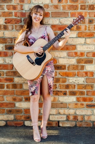 Happy female musician with guitar against brick wall - Australian Stock Image