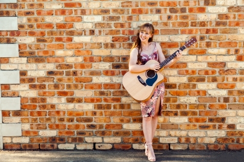 Happy female musician with guitar against brick wall - Australian Stock Image