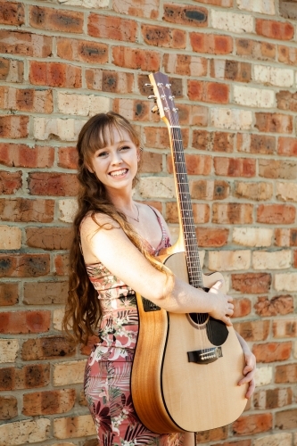 Happy female musician with guitar against brick wall - Australian Stock Image