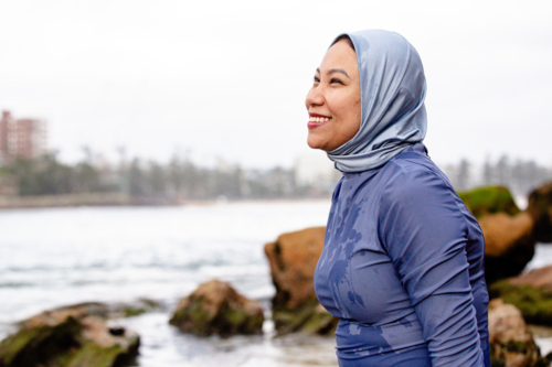 Happy Filipina woman wearing a burkini and light blue headscarf sitting on a rock on the beach - Australian Stock Image
