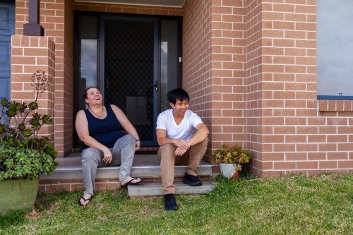 Happy interracial Aussie couple laughing together on front steps of home - Australian Stock Image