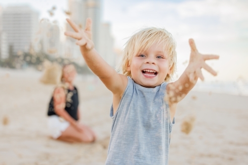 Happy little boy throwing sand on the beach with mother in background - Australian Stock Image