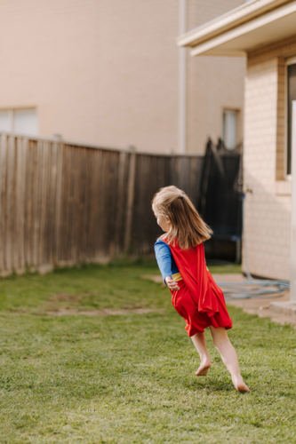 Happy little girl in a superhero costume running around their yard. - Australian Stock Image