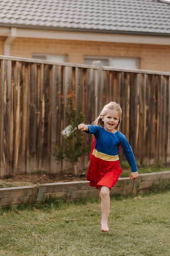 Happy little girl in a superhero costume running around their yard. - Australian Stock Image