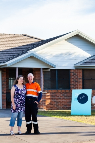 Happy middle aged couple standing infront of house with sold real estate sign - Australian Stock Image