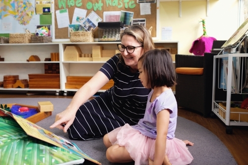 Happy multicultural teacher and child reading a book in kindergarten - Australian Stock Image