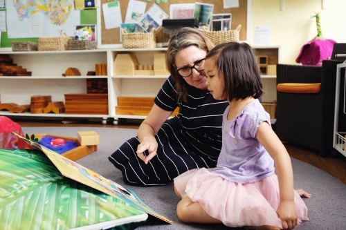 Happy multicultural teacher and child reading a book in kindergarten - Australian Stock Image
