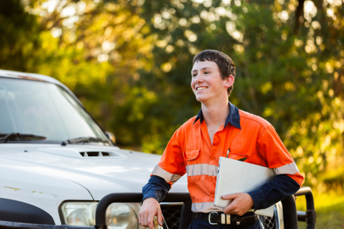 Happy portrait of young Australian man in his teens standing by car holding laptop wearing high-vis - Australian Stock Image