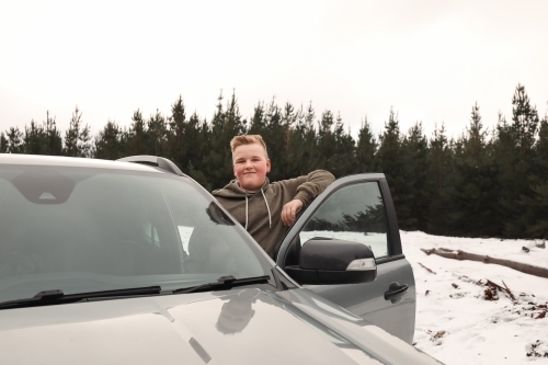 Happy preteen boy standing on side of vehicle smiling in the snow on cold winter drive - Australian Stock Image