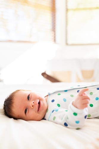 Happy smiling baby lying on parents bed with bassinet in background - Australian Stock Image