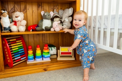 Happy smiling baby standing beside colourful toy shelf in nursery - Australian Stock Image