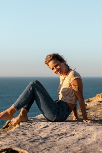 Happy smiling young woman sitting on coastal clifftops at sunrise - Australian Stock Image
