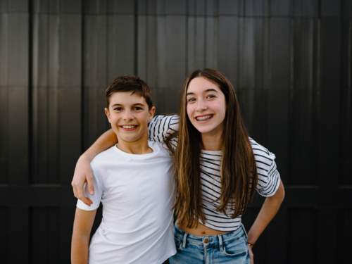 happy teen siblings standing with arms on each others shoulders against black garage door - Australian Stock Image