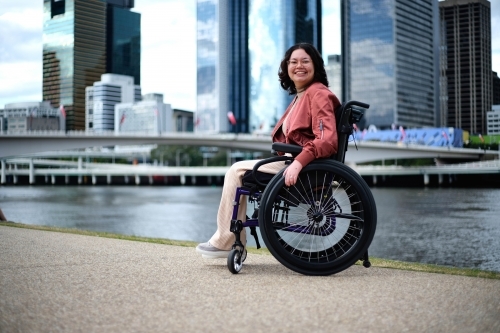 Happy woman in wheelchair in front of river with city in background - Australian Stock Image