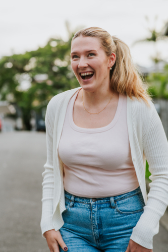 happy woman with a ponytail stands outdoors wearing a white cardigan, pink top, and blue jeans - Australian Stock Image