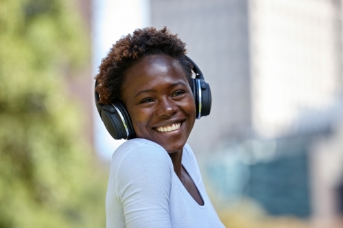 Happy young African woman listening to music wearing wireless headphones in city - Australian Stock Image