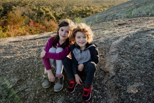 Happy young brother and sister sitting together on a rocky ledge in the Australian bush at sunset