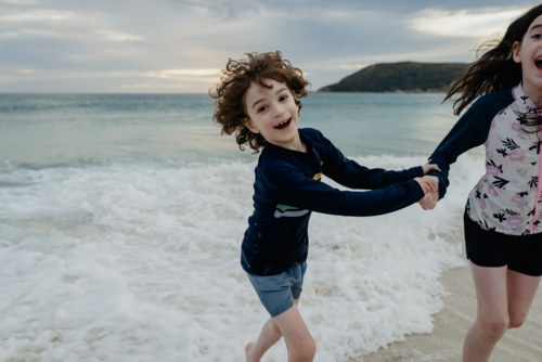 Happy young brother and sister (young boy & girl) holding hands running in the surf at the beach - Australian Stock Image
