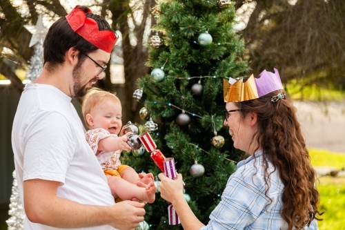 Happy young family - couple with baby celebrating Christmas with silly paper hat crowns and bon bons - Australian Stock Image
