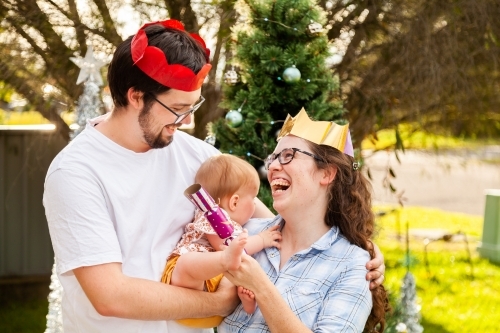 Happy young family - couple with baby laughing together with Christmas bon bon cracker - Australian Stock Image