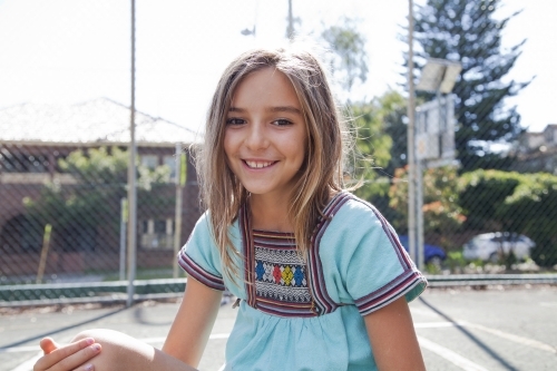 Happy young girl sitting inside court surrounded by chain-link fence - Australian Stock Image
