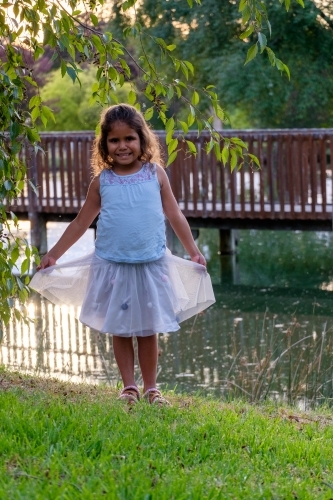 happy young girl standing on grass beside dam in park holding skirt - Australian Stock Image