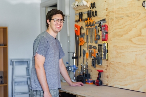 happy young man in home shed leaning on workbench with tools on board behind him - Australian Stock Image