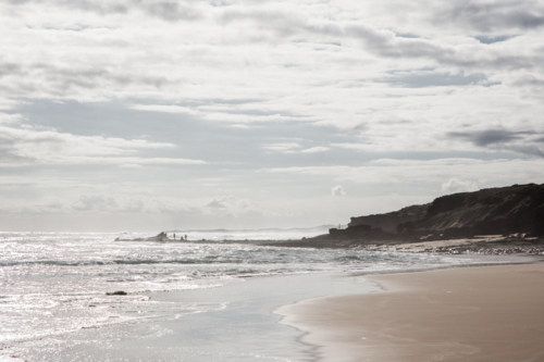 Hazy, late afternoon beach with people on rocks in the distance - Australian Stock Image