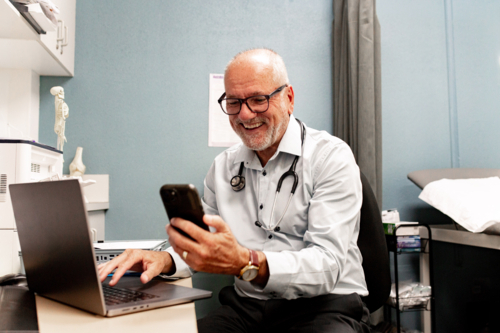 Healthcare professional at desk smiling while working on his laptop and checking smartphone - Australian Stock Image