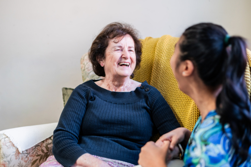 Healthcare worker engaged in a conversation with an elderly woman seated on patterned armchair - Australian Stock Image