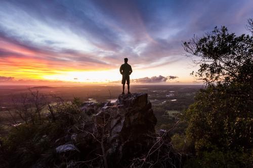 Hiker standing atop a rock formation watching sunrise from Mount Tibrogargan - Australian Stock Image