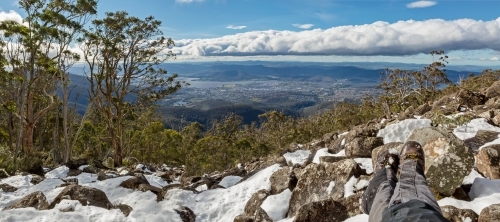 hiker's gaitered legs and boots relaxed on snowy rocks with view over Hobart