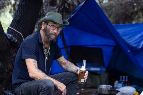 homeless man sitting at his secluded camping spot under trees drinking beer - Australian Stock Image