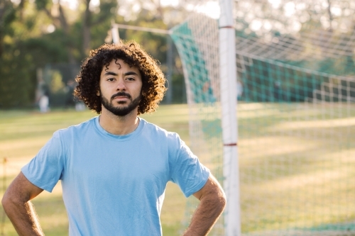 horizontal half body shot of a man with curly hair with arms in the side with goal in the background