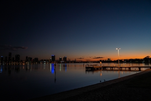 horizontal shot of a jetty with lamp post during sunset with buildings reflected by the water - Australian Stock Image