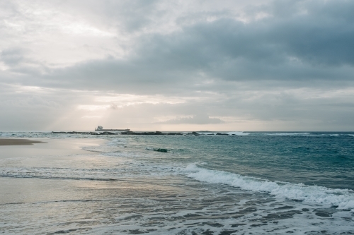 horizontal shot of a beach with waves, rocks and ship at a distance on a cloudy day - Australian Stock Image