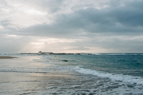 horizontal shot of a beach with waves, rocks and ship at a distance on a cloudy day